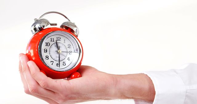 A Caucasian hand, belonging to an adult, holds a red alarm clock prominently in the foreground, with copy space. Time management and punctuality are emphasized in this image, suggesting the importance of being on time or the concept of an impending deadline.