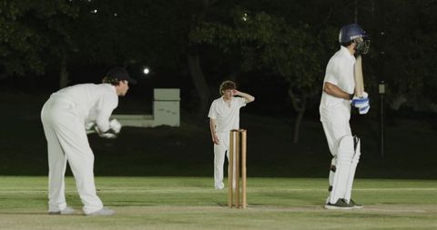 Night cricket action: bowling, wicketkeeping and batting on floodlit grass pitch showing teamwork