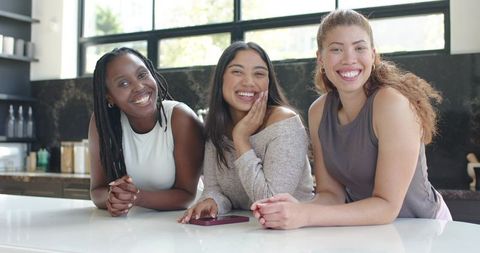 Diverse friends leaning on kitchen island smiling while sharing smartphone and casual chat