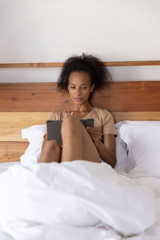 African american woman relaxing in bed using tablet under white duvet and wooden headboard