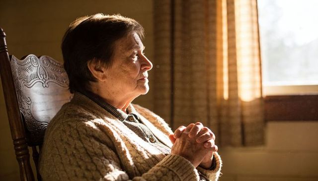 Senior woman sitting in carved chair wearing beige cardigan, gazing through sunlit window
