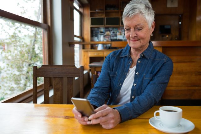Senior woman sitting at a wooden table in a cozy café, using a mobile phone while enjoying a cup of coffee. Ideal for illustrating themes of modern technology use among the elderly, relaxed lifestyle, and casual café settings.