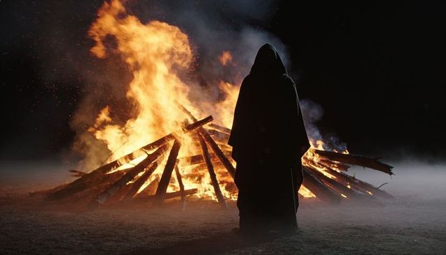 Hooded figure watching bonfire at night, smoky ritual silhouette and mystery