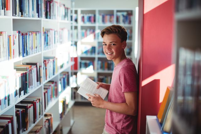 Portrait of happy schoolboy reading book in library at school