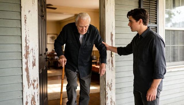 Young man helping elderly man with cane stepping through weathered porch doorway