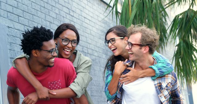 Young adults of diverse ethnic backgrounds are seen hugging and smiling outdoors. The casual attire and background of green plants and a brick wall suggest a laid-back, friendly atmosphere. Perfect for use in advertisements showcasing diversity, friendship, unity, or any promotional material aimed at young adults.