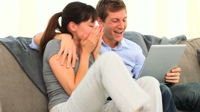 Young couple sitting on a sofa together, expressing excitement and joy while looking at a tablet. Suitable for themes related to online gaming victories, internet success stories, digital happiness, or fintech marketing.