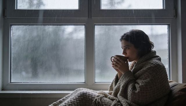 Woman wrapped in knitted blanket sipping warm mug on rainy window seat, cozy introspection