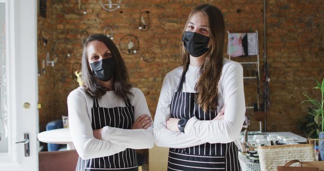 Two female cafeteria staff members wearing striped aprons and protective black masks stand indoors with a brick wall behind them. They appear ready to serve and attentive to safety protocols, embodying professionalism and teamwork. This is perfect for illustrating themes of customer service, hospitality industry, health protocols, small businesses, and teamwork.
