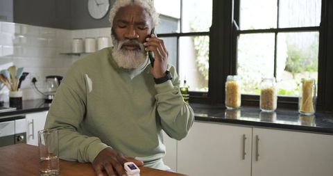 Senior man monitoring pulse with oximeter while on phone in kitchen