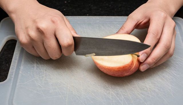 Hands slicing red apple with chef knife on plastic cutting board for recipe prep