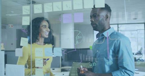 Two coworkers brainstorming on glass wall with sticky notes, markers and flowchart diagrams