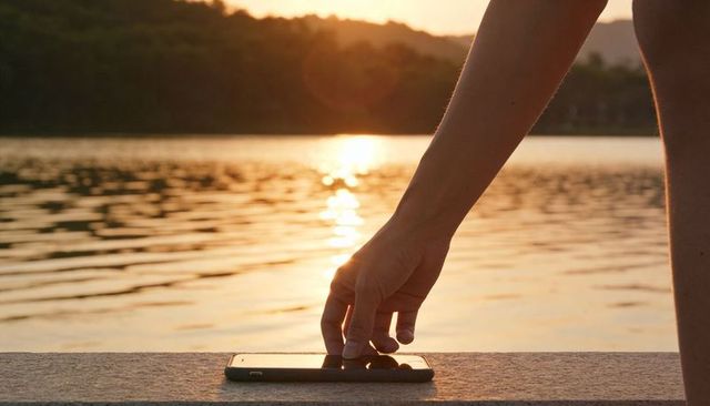 Reaching hand touching smartphone on stone ledge while sun reflects on calm lake