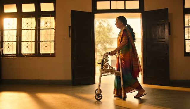 Senior woman walking with rollator in sunlit foyer wearing colorful silk saree