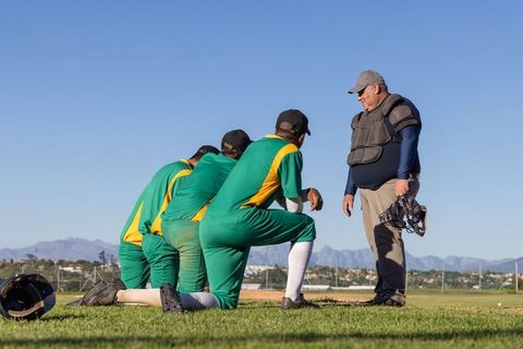 Umpire Holding Mask Coaching Players Kneeling at Home Plate on Sunlit Baseball Field