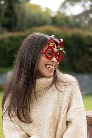 Young woman laughing wearing red glitter christmas tree glasses and cream knit turtleneck