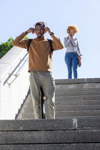 African american man adjusting headphones with skateboard while woman talking on phone on stairs