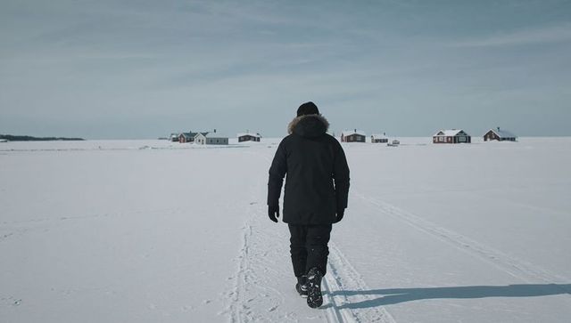 Solitary man walking across frozen snowfield toward distant cabins under overcast sky