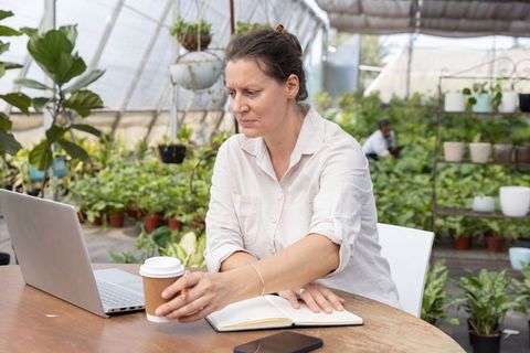 Mature woman working in greenhouse workspace reaching for coffee while using laptop
