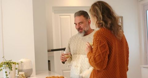 Middle-aged couple sharing wine at kitchen counter holding glasses with pastry cozy sweaters