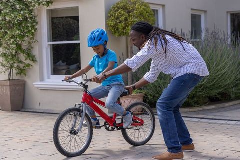 African american father teaching son to ride red bike wearing blue helmet on home driveway