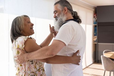 Mature couple dancing and embracing in sunlit living room near window blinds