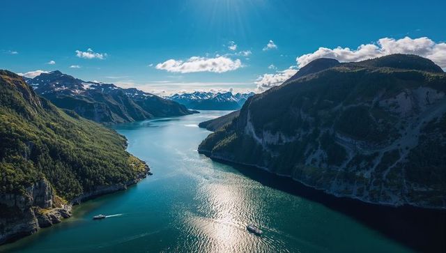 Sunlit alpine fjord with turquoise water and snow-capped peaks, boats leaving wakes