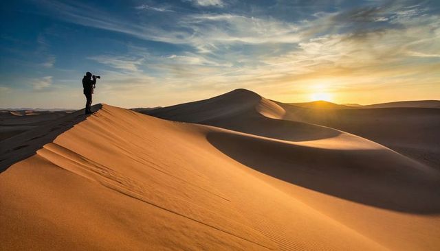 Photographer capturing sunset over rolling desert dunes with telephoto lens and footprints