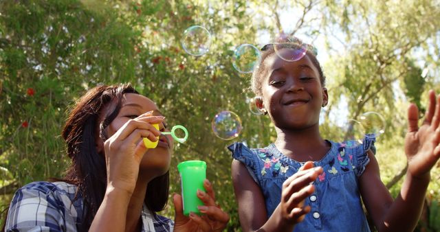 A young African American woman and a girl are enjoying a sunny day outdoors blowing bubbles, with copy space. Their joyful activity captures a moment of carefree happiness and bonding.