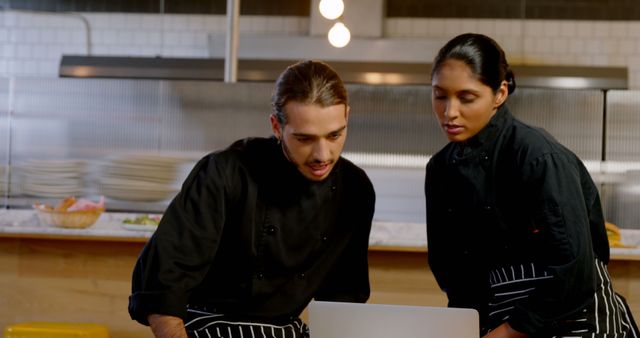 Two chefs, one with long hair and the other with dark hair, work on a laptop in a commercial kitchen. They are both dressed in black uniforms and appear to be collaborating on a project, indicating the use of technology in modern culinary practices. Ideal for illustrating themes of teamwork, the integration of technology in cooking, and professional kitchens.