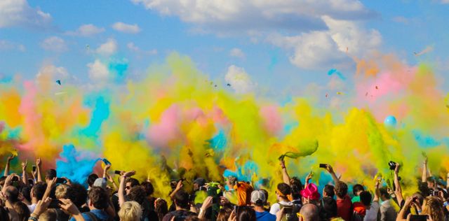 This dynamic image captures a large crowd celebrating with colorful powder clouds under a bright blue sky. Perfect for depicting joyful events like music festivals, cultural celebrations, or outdoor activities. Ideal for use in promotional materials, event advertisements, and social media content showcasing fun and vibrant gatherings.