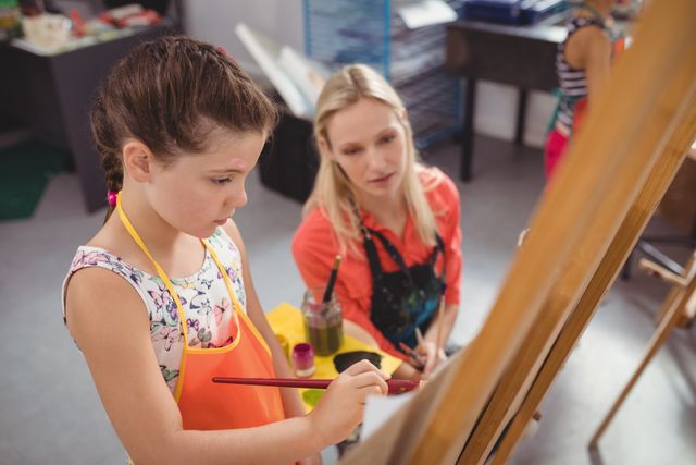 Teacher assisting young girl in an art class, providing guidance and support as she paints on an easel. Ideal for use in educational materials, school brochures, art class promotions, and articles about creative learning and child development.