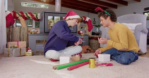 Diverse Gay Couple Wrapping Christmas Gifts by Cozy Fireplace