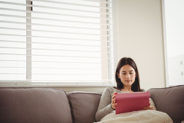 Young woman sitting on a comfortable sofa at home, using a tablet. Ideal for themes related to technology, home lifestyle, relaxation, and modern living. Perfect for articles, blogs, or advertisements focusing on digital devices, home comfort, or leisure activities.