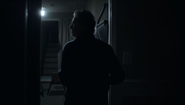 Backlit silhouette of man standing in dark doorway with illuminated staircase and corridor