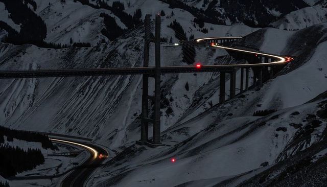 Snowy alpine viaduct glowing at night with curving light trails and cable-stayed bridge