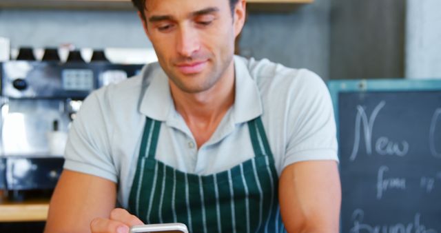 Young male barista in striped apron using smartphone in modern cafe. Ideal for depicting modern technology in small businesses, featuring communication, work-life balance, and casual work environments in coffee shops. Useful for articles related to barista life, coffee culture, small business management, and the integration of digital tools in service industries.