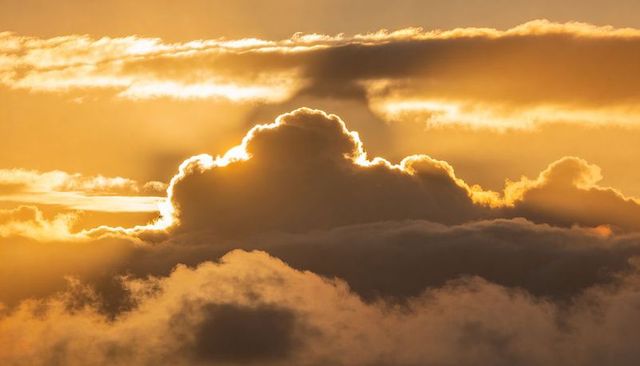 Golden rim-lit cumulus cloud glowing at sunset with layered mist and dramatic sunbeam sky