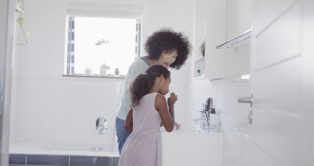 African American mother helping young daughter brush her teeth in a bright, modern bathroom with white decor and natural light coming through the window. Suitable for articles or advertisements related to parenting, family life, daily routines, oral hygiene practices, and child care.