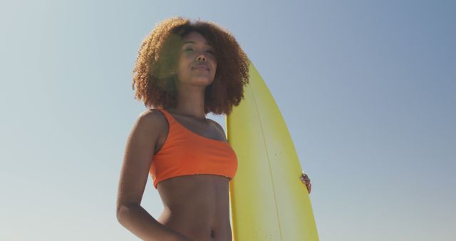 Happy biracial woman holding surfboard standing on sunny beach and looking out to sea. Summer, hobbies, surfing and vacations.