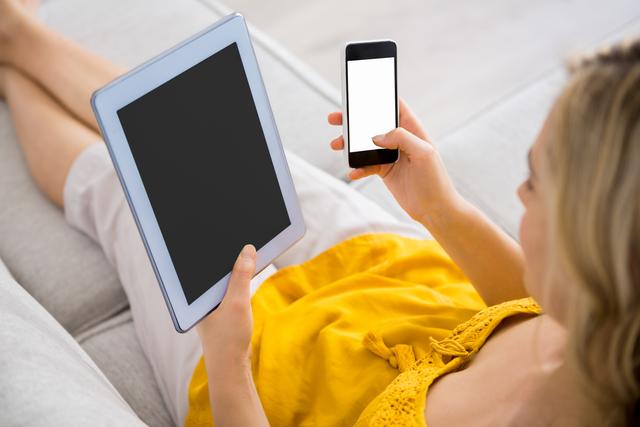 Woman sitting on couch using mobile phone and tablet, ideal for illustrating modern lifestyle, technology use, and digital communication. Perfect for blogs, articles, and advertisements related to tech, home living, and connectivity.