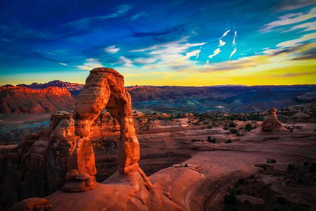 Photo showcases the iconic Delicate Arch in Arches National Park at sunset. Sky is filled with vibrant colors contrasting with the red sandstone. Ideal for promoting tourism in Utah, travel blogs, outdoor adventure advertisements, and nature magazines.