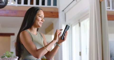 Smiling woman tapping smartphone in sunlit loft near sliding glass doors with smart home app