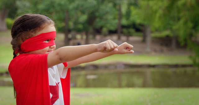 A young girl wearing a red superhero cape and mask is playing outdoors in a park. She stretches her arms forward, pretending to fly. This image is ideal for promoting creative play, childhood imagination, or children's apparel. It can also be used in educational materials that encourage outdoor activities and adventure, or in advertisements for toys and games.