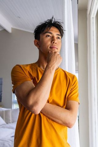 Asian man contemplating at sunlit window wearing mustard tee, minimal bedroom lifestyle