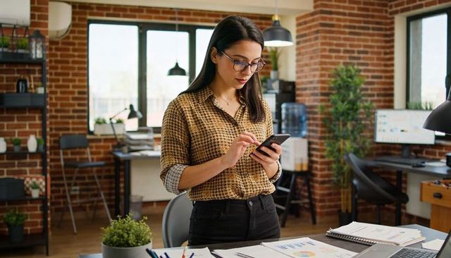Professional checking smartphone in modern brick office workspace with charts and natural light