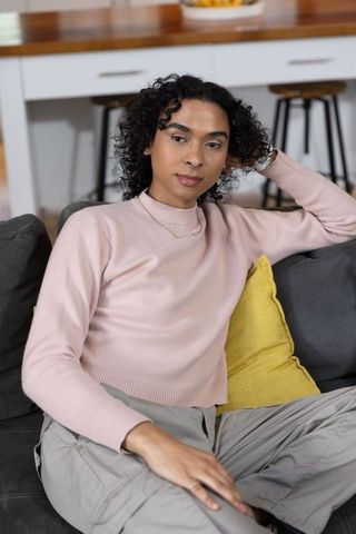 Non-binary person relaxing on sofa wearing pink sweater with yellow cushion, modern kitchen
