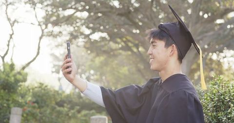 Asian graduate taking selfie in cap and gown on sunlit campus with tassel and smile