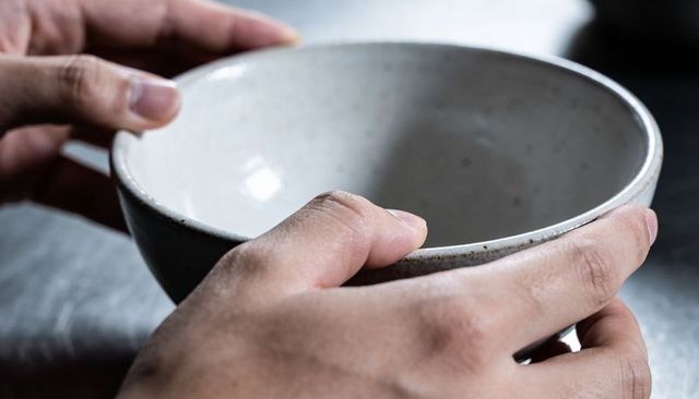 Cradling hands holding speckled ceramic bowl on textured grey tabletop minimalist closeup