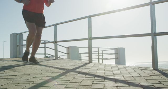 Lower body of man in athletic wear jogging on a paved walkway near the waterfront on a sunny day. Legs are in motion, casting shadows, with sunlight creating flares. Ideal for use in fitness promotions, advertisements for activewear, healthy lifestyle campaigns, and outdoor activity blogs.